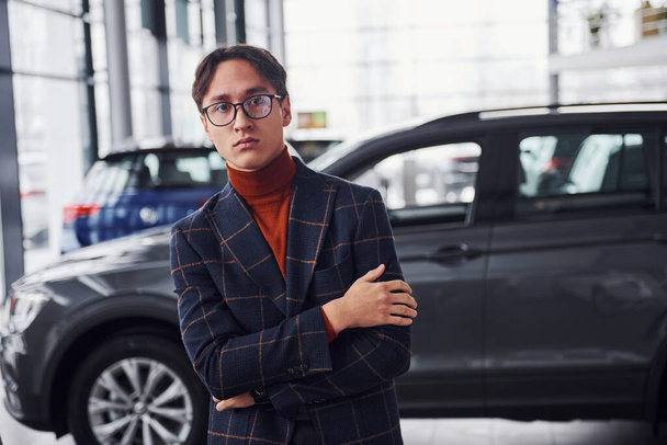 Posing for the camera. Young business man in luxury suit and formal clothes is indoors near the car. - 写真・画像