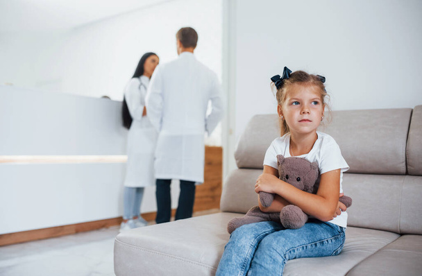 Doctors behind. Cute little girl with teddy bear in hands sits in waiting room of hospital. - Foto, afbeelding