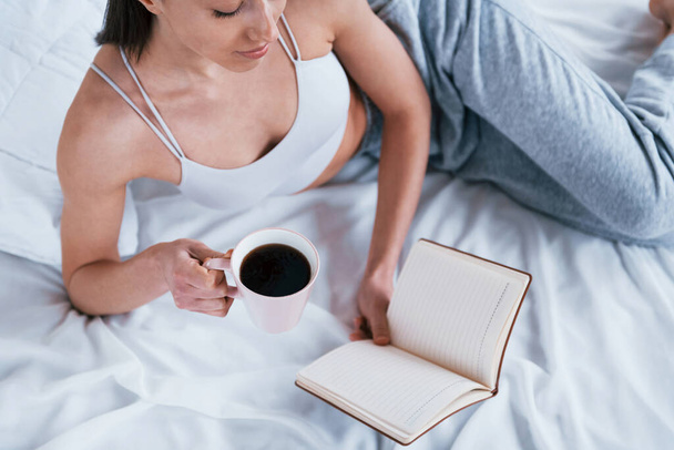 Femme souriante positive en vêtements de sport couchée dans le studio avec livre et tasse de boisson dans les mains. - Photo, image