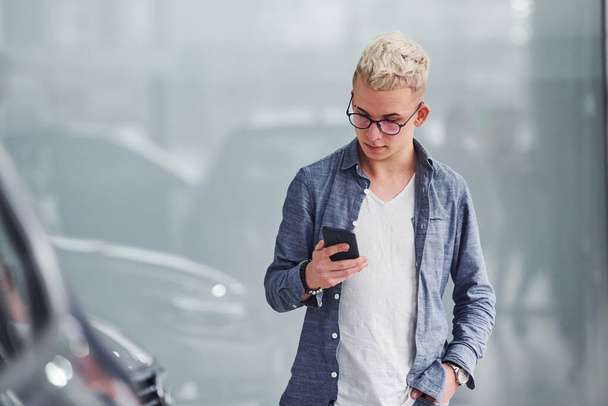 Young hipster guy in nice clothes stands indoors against grey background near modern car. - Foto, immagini