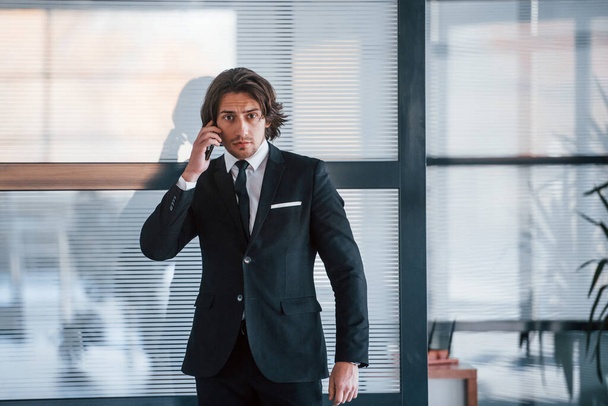Talking by the phone in the office. Portrait of handsome young businessman in black suit and tie. - Fotografie, Obrázek