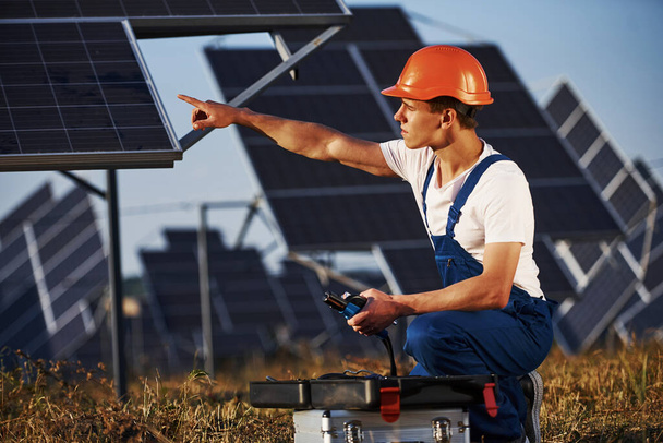 Using special equipment. Male worker in blue uniform outdoors with solar batteries at sunny day. - Foto, Imagen