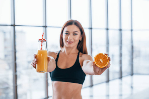 Sportive woman with nice body stands indoors with bottle of orange juice and fruit in hands. - Foto, Imagem