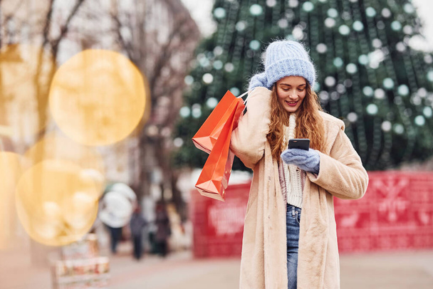 Young girl in warm clothes with shopping bags in hand have a walk outdoors in the city and holding phone. - Photo, Image