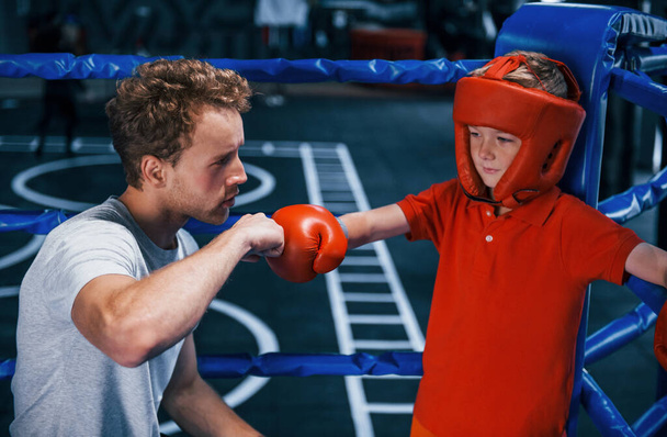 Young boxing coach is helping little boy in protective wear on the ring between the rounds. - Fotó, kép