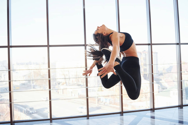 Young sporty woman in sportswear jumping and doing athletic tricks against window in the air. - 写真・画像