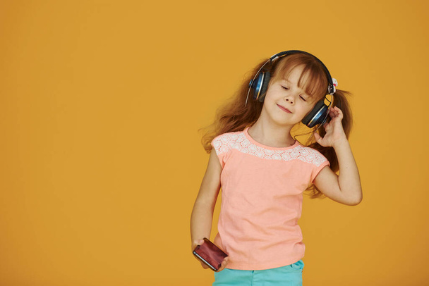 Portrait of cute little girl in headphones and with phone in the studio against yellow background. - Foto, Imagem