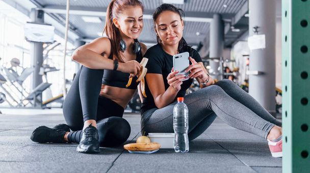 Two female friends in sportive clothes is in the gym earing fruits and using phone. - Photo, Image