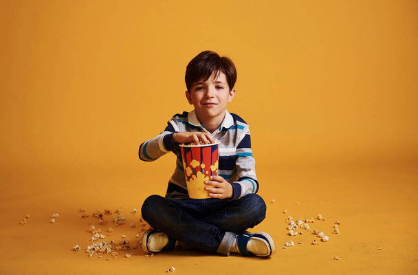 słodkie mały chłopiec siedzi i ogląda film i je popcorn przeciw żółty ściana. - Zdjęcie, obraz