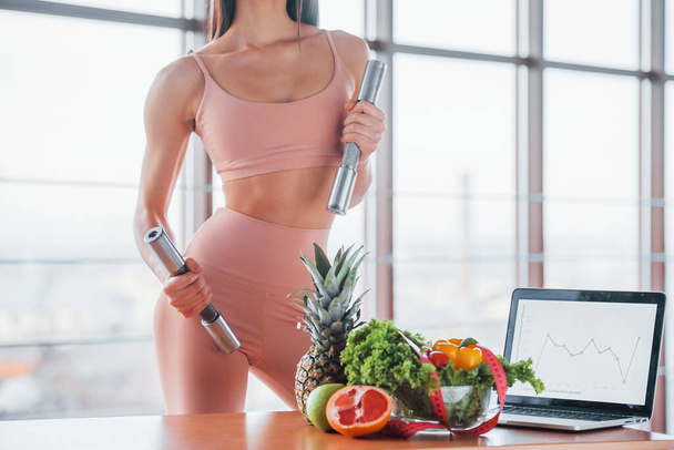 Female athlete in sportive wear holds dumbbells and standing near table with laptop and healthy food. - Foto, Imagem