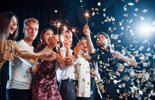 Having fun with sparklers. Confetti is in the air. Group of cheerful friends celebrating new year indoors with drinks in hands. - Фото, изображение