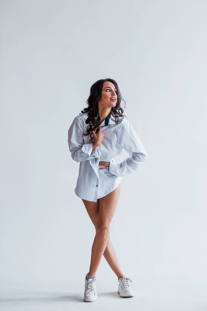 Cheerful young brunette in white coat and with sexy legs stands in the studio. - Foto, Bild