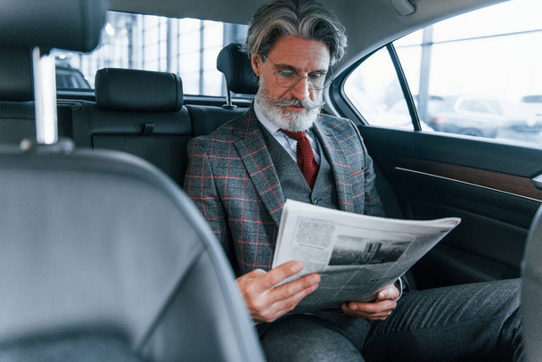 Modern stylish senior man with grey hair and mustache reading newspaper inside of the car. - Fotó, kép