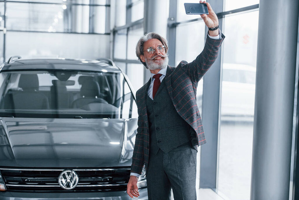 Positive senior man with grey hair taking selfie against modern car indoors. - Foto, Imagem