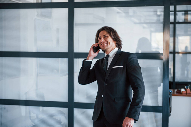 Talking by the phone in the office. Portrait of handsome young businessman in black suit and tie. - Zdjęcie, obraz