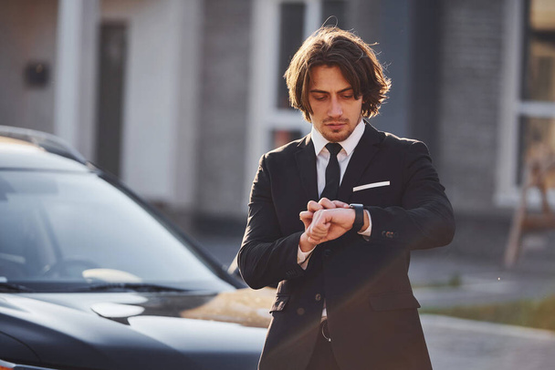 Portrait of handsome young businessman in black suit and tie outdoors near modern car in the city. - Foto, immagini