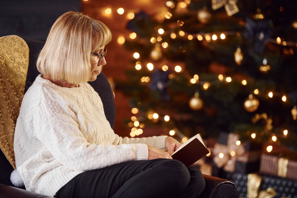 Old woman sitting on the chair with book in festive New year room with christmas decorations. - Photo, image