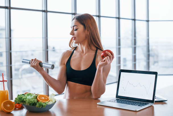 Sportive woman with nice body stands indoors with dumbbell and fresh apple in hands near table with laptop. - Photo, image