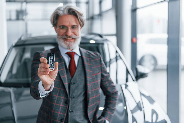 Senior businessman in suit and tie with gray hair and beard standing indoors near car with keys in hand. - Foto, Imagem