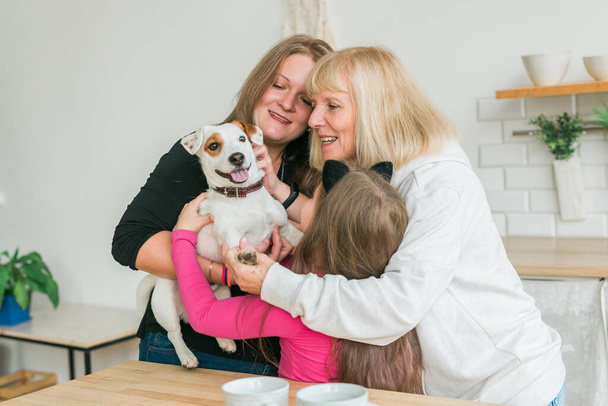 Bonne petite-fille et grand-mère et fille s'amuser avec Jack Russell terrier chien. Grand-mère embrasse son petit-enfant à la maison. Relation, famille et concept de trois générations. - Photo, image