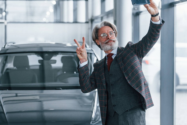 Positive senior man with grey hair taking selfie against modern car indoors. - Фото, изображение