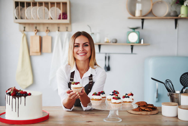 Positive blonde with her homemade delicious cookies and cake in the kitchen. - Foto, afbeelding