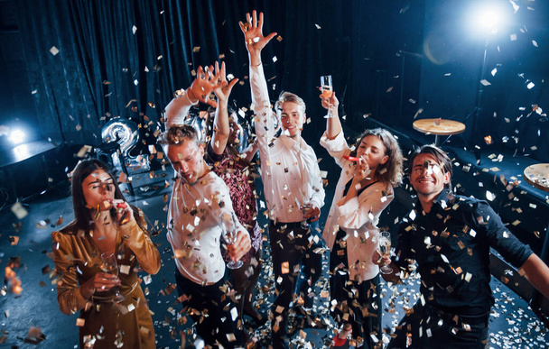 Confetti is in the air. Group of cheerful friends celebrating new year indoors with drinks in hands. - Fotoğraf, Görsel