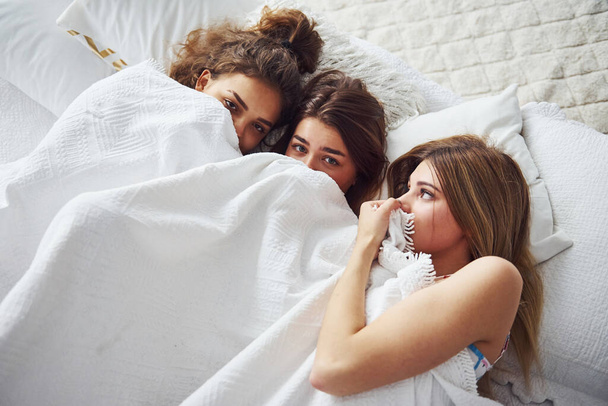 Lying on the bed under the blanket together. Happy female friends having good time at pajama party in the bedroom. - Photo, Image