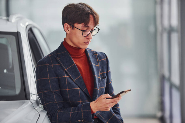 Hipster in formal wear and in eyewear standing near white modern car indoors. - 写真・画像