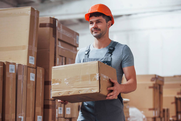 Young male worker in uniform is in the warehouse with box in hands. - Foto, afbeelding