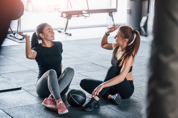 Two young girls in sportive clothes is in the gym together at daytime sitting on the floor with ball and giving high five. - Fotoğraf, Görsel