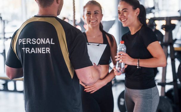 Two young girls in sportive clothes is in the gym together at daytime with their male personal trainer. - Foto, afbeelding