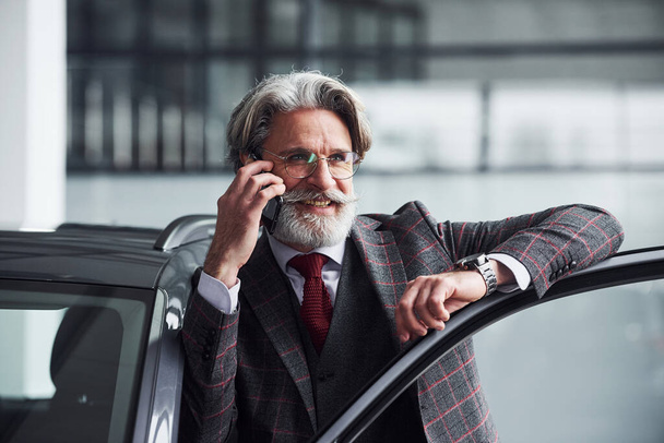 Senior businessman in suit and tie with gray hair and beard is near car talking by the phone and smiling. - Photo, image