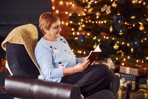 Vanha nainen istuu tuolilla kirjan kanssa juhlava uudenvuoden huone joulukoristeilla. - Valokuva, kuva