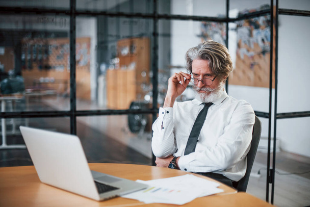 Verveelde volwassen zakenman met grijs haar en baard in formele kleding is in het kantoor tegen laptop. - Foto, afbeelding
