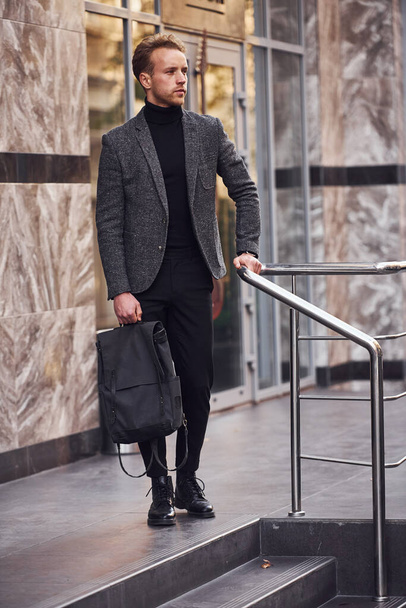 Man in elegant formal wear and with bag is outside against modern building. - Photo, image