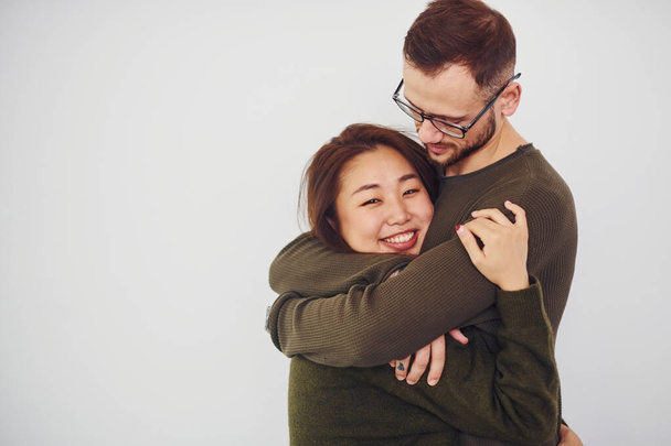 Happy multi ethnic couple in casual clothes embracing each other indoors in the studio. Caucasian guy with asian girlfriend. - 写真・画像