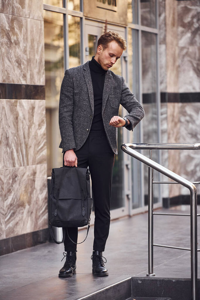 Man in elegant formal wear and with bag is outside against modern building. - Фото, зображення