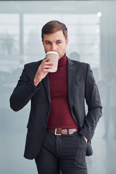 Young business man in luxury suit and formal clothes wth cup of drink is indoors in the office. - Photo, image