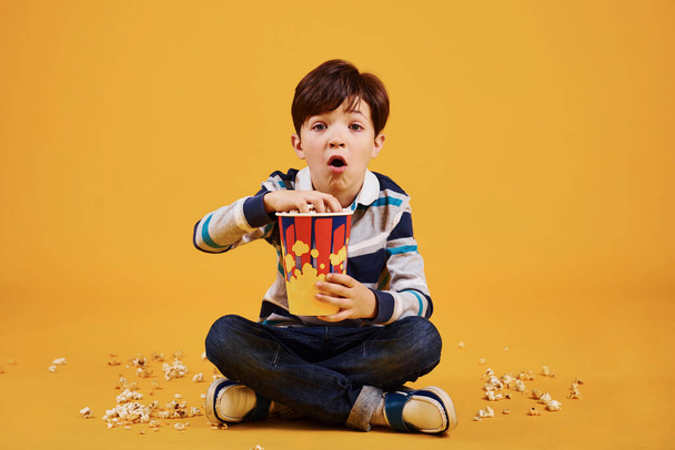 słodkie mały chłopiec siedzi i ogląda film i je popcorn przeciw żółty ściana. - Zdjęcie, obraz