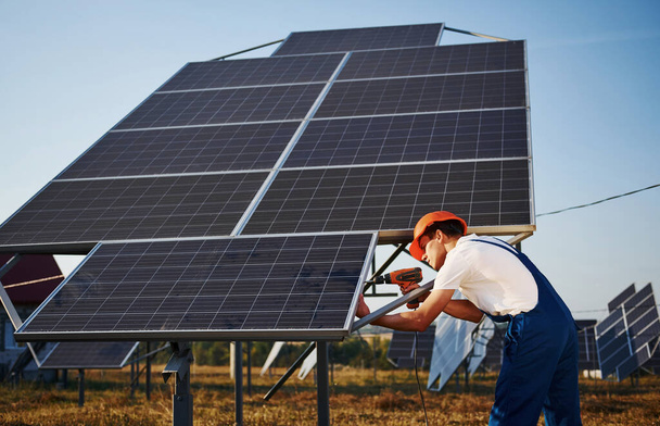 Usando destornillador inalámbrico. Hombre trabajador en uniforme azul al aire libre con baterías solares en el día soleado. - Foto, Imagen