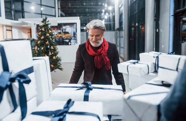 Stylish old man with grey hair and mustache packing presents in gift boxes into car. Christmas tree at background. - Zdjęcie, obraz