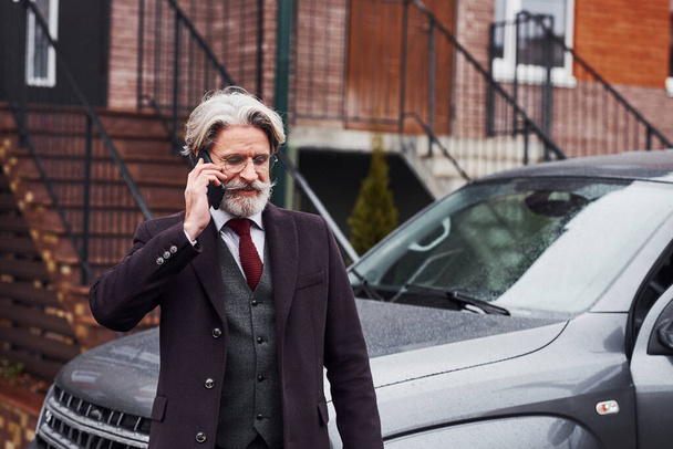 Fashionable senior man with gray hair and beard is outdoors on the street near his car have conversation by the phone. - Photo, image