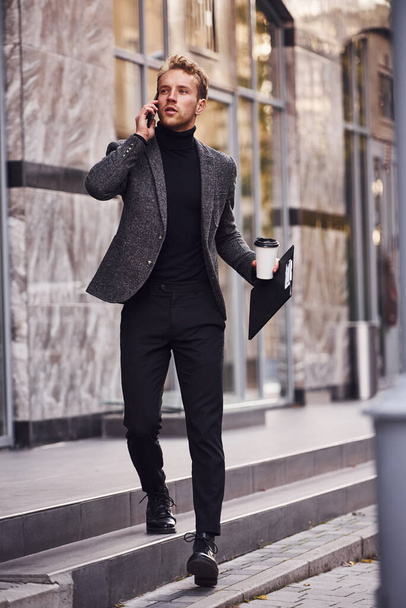 Man in elegant formal wear with cup of drink and notepad in hands is outside against modern building have a conversation by the phone. - Фото, изображение