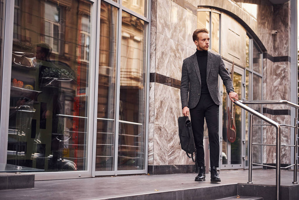 Man in elegant formal wear and with bag is outside against modern building. - Foto, Imagen