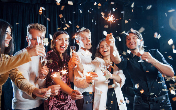 Having fun with sparklers. Confetti is in the air. Group of cheerful friends celebrating new year indoors with drinks in hands. - Photo, Image