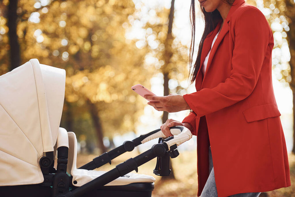 With phone in hand. Mother in red coat have a walk with her kid in the pram in the park at autumn time. - Foto, Imagem