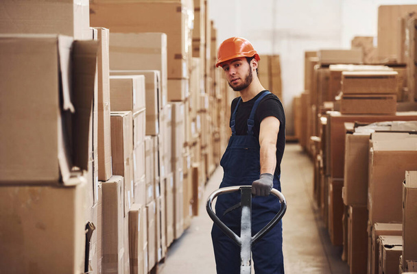 Young male worker in uniform is in the warehouse pushing pallet truck. - 写真・画像
