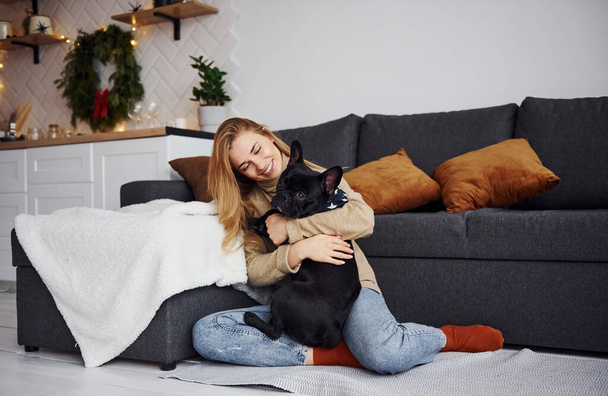 Cute young girl in casual clothes sitting near the sofa indoors at home and embracing the dog. - Fotó, kép
