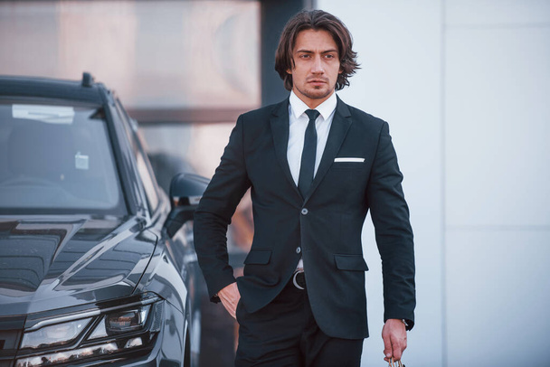 Portrait of handsome young businessman in black suit and tie outdoors near modern car. - Photo, Image